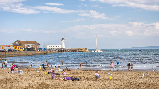 Scarborough, UK. August 13, 2022.   People enjoy leisure time on a beach.  There is a lighthouse and pier beyond with a yacht entering harbour. A blue sky with cloud is above.