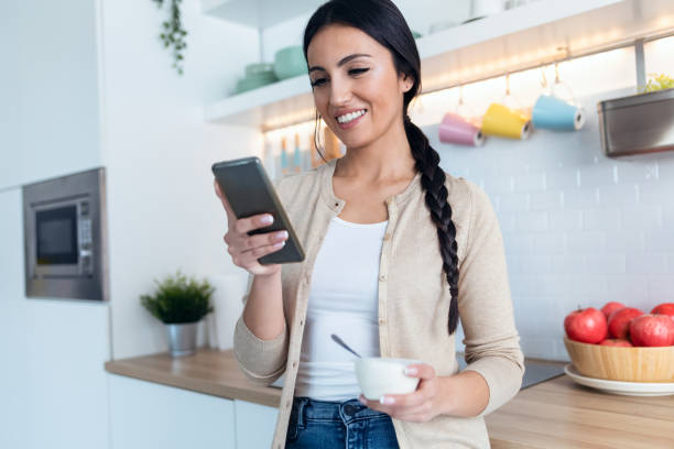istock Pretty woman sending messages with mobile phone while eating yogurt int he kitchen at home. 1418492080