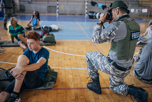 Diverse group of people, soldiers on humanitarian aid to civilians in school gymnasium, after natural disaster happened in city. Army press is taking photographs of people.