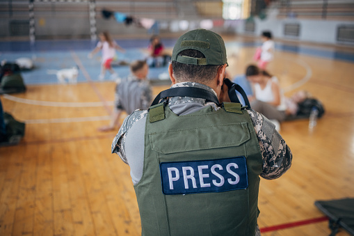 Diverse group of people, soldiers on humanitarian aid to civilians in school gymnasium, after natural disaster happened in city. Army press is taking photographs of people.