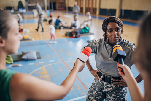 Diverse group of people, soldiers on humanitarian aid to civilians in school gymnasium, after natural disaster happened in city. Television press talking to a soldier.