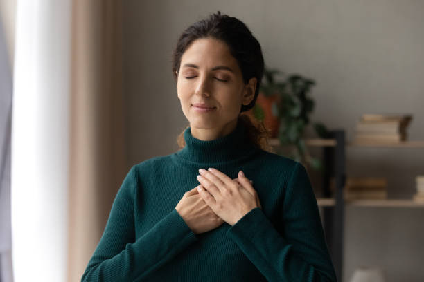 Peaceful young woman feeling thankful at home. Head shot peaceful young attractive woman holding folded hands on chest, praying waiting for miracle, feeling thankful indoors. Sincere happy female volunteer showing kindness or expressing gratitude. begging social issue stock pictures, royalty-free photos & images