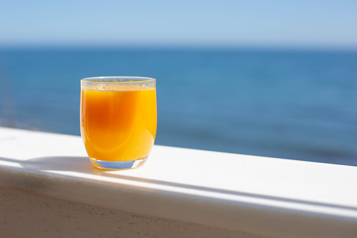 A glass of orange juice is  on the fence of the terrace against the background of the blue sea. Free space for text.