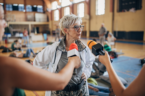 Diverse group of people, soldiers on humanitarian aid to civilians in school gymnasium, after natural disaster happened in city. Television press talking to a female army doctor.