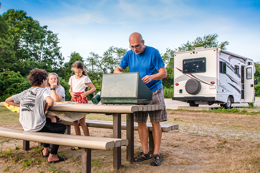 Mature man cooking on outdoor propane stove on a picnic table during summer day
The family is sitting besides him with the motorhome behind