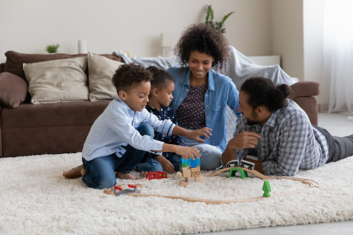 Happy young African parents teaching two little son and daughter to construct city road, highway, playing creative game with toy building blocks on white soft carpeted floor. Family leisure concept