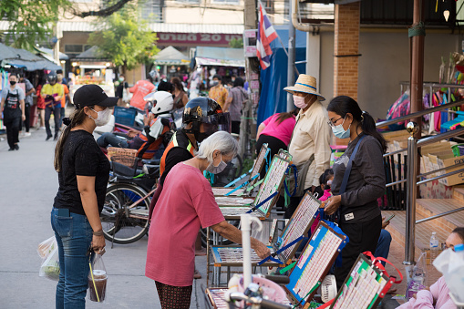Thai senior woman is buying lottery ticket from lottery vendors with bicycles. Scene is close to a local market in Bangkok Chatuchak. Vendors are at right side. A man is waiting behind senior woman. People are wearing protective face masks