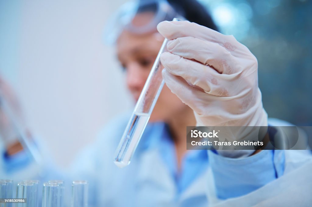 Focus on a test tube with chemical substance in the gloved hand of a pharmacologist working in a scientific laboratory Selective focus on a test tube with a chemical in the hand of a blurred scientist, pharmacologist working on a new drug in a research laboratory. Pharmacy and medicine. Clinical Study Adults Only Stock Photo