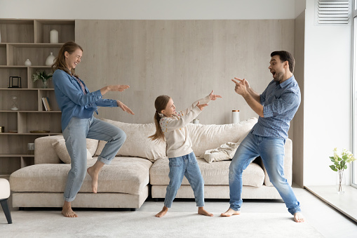 Excited funny parents and sweet happy daughter kid playing western, making gun hand gunshots, dancing to hip hop music in living room, laughing, having fun, enjoying active games together