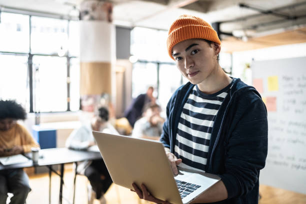 portret młodego mężczyzny trzymającego laptopa w klasie lub małej firmie - computer programmer laptop men nerd zdjęcia i obrazy z banku zdjęć