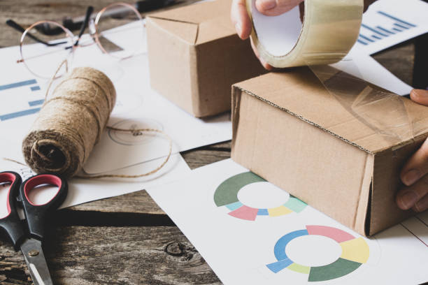 Women's online store, small business owner, entrepreneur, packaging box and preparing a parcel on the table for shipment. Dropshipping delivery concept in e-commerce. Product packaging, bar graph and revenue totals tienda en línea para mujeres, propietaria de una pequeña empresa, empresaria, caja de embalaje y preparación de un paquete sobre la mesa para el envío. concepto de entrega dropshipping en e-commerce. embalaje del producto, gráfico de barras