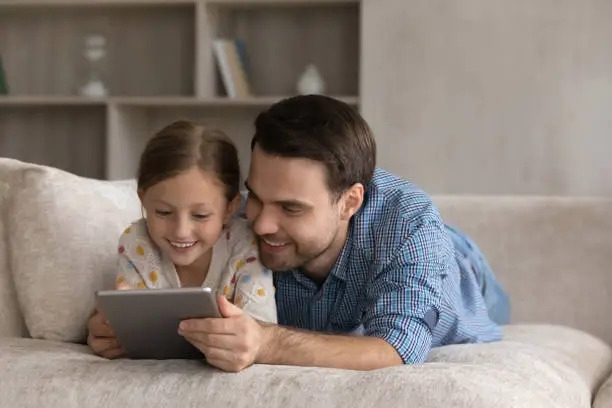 Cheerful millennial father hugging sweet gen Z daughter girl lying on sofa at home, using learning application, tablet computer, playing online videogame on digital gadget, reading ebook on internet