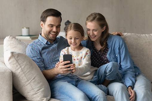 Happy couple of parents and sweet daughter kid resting on comfortable cough in living room, using mobile phone, talking on video call, shopping on internet, using online ecommerce app