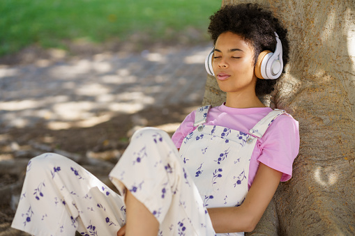 Calm African American female with closed eyes listening to music in wireless headphones while resting near tree trunk in park
