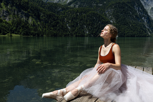 Young ballerina sits on a wooden stage by the lake. High quality photo