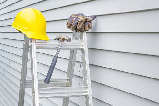Hard hat, work gloves and hammer on stepladder in front of house siding