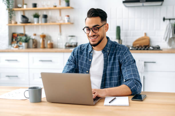 istock Handsome positive indian or arabian male entrepreneur, freelancer, working remotely while sitting at home in the kitchen, using laptop, typing on keyboard, develop creative successful project, smiling 1418580403