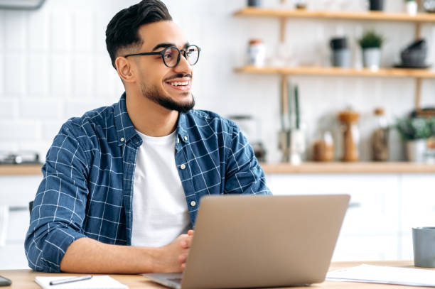 istock Handsome positive stylish indian or arabian young man with glasses, programmer, freelancer, working remotely, sitting at home in the kitchen with laptop, looking to the side, dreaming, smile happily 1418580413