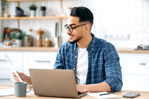 istock Job with documents. Smart successful arabian or indian guy with glasses, freelancer or office worker working remotely, sit at a desk at in s kitchen, working on a project, analyzing documents, smiling 1418580425