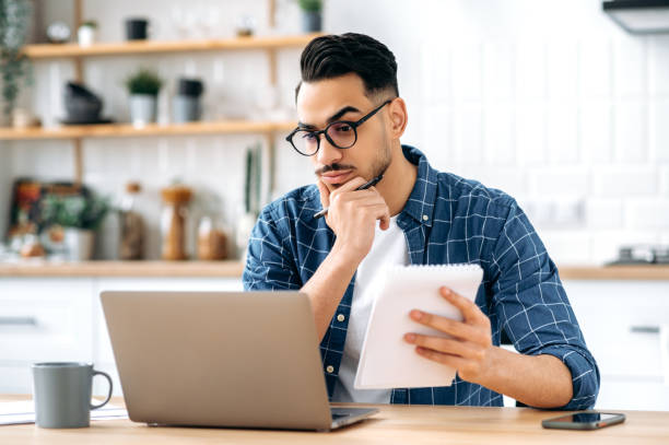 istock Smart focused modern stylish arabian or indian young man, working on a project while sitting at home and using a laptop, looking at the screen thoughtfully, planning a strategy, analyzing risks 1418580430
