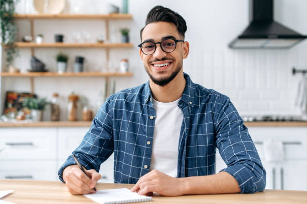 istock Happy positive Arabian or Indian modern guy, clever student or freelancer, with glasses, listening to an online webinar, lecture, writing down information, looking at the camera, smiling friendly 1418580496