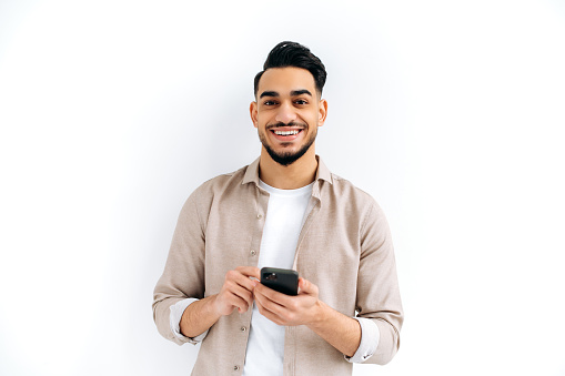 Handsome positive Arabian or Indian modern guy, holding smartphone in his hands, messaging, looking excitedly at the camera, standing over isolated white background, smiling