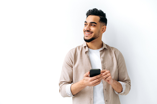 Confident positive handsome indian or arabian young man, holding smartphone in hand, chatting online, browsing internet, looking happily to the side, standing on isolated white background, smiling