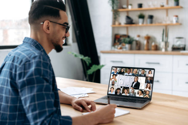 istock Online education. View over the shoulder of a mixed race guy taking notes in a notebook, at a laptop screen with a teacher and a group of multiracial students. E-learning, webinar, online lecture 1418582664
