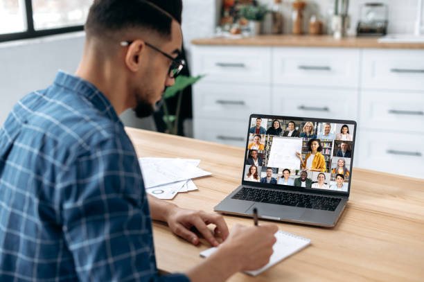 istock Distance learning, online course. View over the shoulder of a guy taking notes in a notebook, at a laptop screen with a teacher and a group of multiracial students. E-learning, webinar, online lecture 1418582680