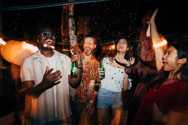 A group of friends is celebrating with confetti at a nighttime rooftop party. A cheerful group of multiracial friends is celebrating and having fun with confetti at the open-air summer rooftop nightclub. They are having a blast, dancing and drinking beer. nightclub stock pictures, royalty-free photos & images