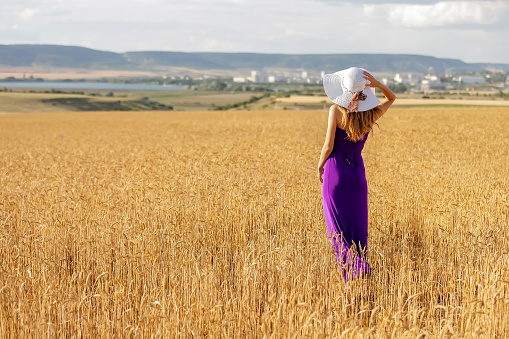 Happy young woman holding hat, walking in the golden wheat field. Summer nature. Sunset light.