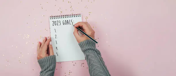 Photo of Woman's hand writing 2023 goals in note pad on pink background. Top view. Concept of New Year's plans, goals and actions