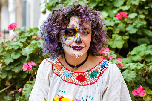 portrait of beautiful young latin woman smiling and looking at the camera, with La Calavera Catrina makeup sitting outdoors with plants and flowers in the background