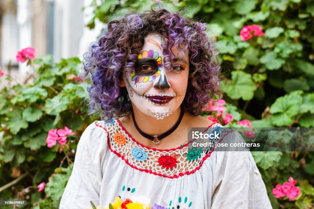 portrait latin young woman with La Calavera Catrina makeup with plants and flowers in the background portrait of beautiful young latin woman smiling and looking at the camera, with La Calavera Catrina makeup sitting outdoors with plants and flowers in the background Latin American and Hispanic Ethnicity Stock Photo