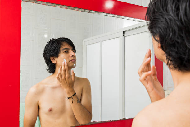 istock young man putting cream on his face while looking at himself through a mirror 1418614365