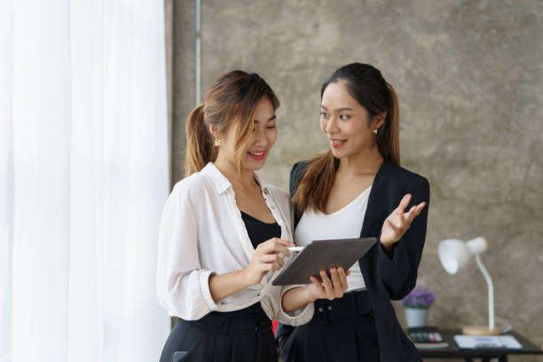 istock Two beautiful Asian businesswomen standing using digital tablet consulting and analyzing information in office work. 1418641432
