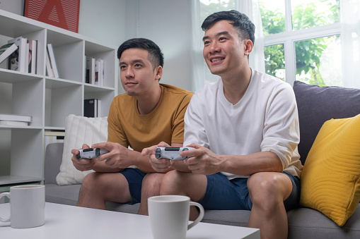 9267-9276
Young Asian gay couple playing video games on a couch and having fun at home. LGBT couple looking happy while spending time together at home.