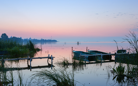 Gentle pink and blue dawn over the calm misty lake before sunrise. Moored boats at the old wooden pier near the overgrown bank. Travel concept. Lake Nero, Rostov Veliky, Russia.