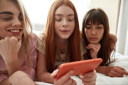 Young european girlfriends watching mobile phone in hand of red haired girl on bed during girlish pajama party at home. Zoomer girls spending time together. Friendship. Rest, entertainment and leisure