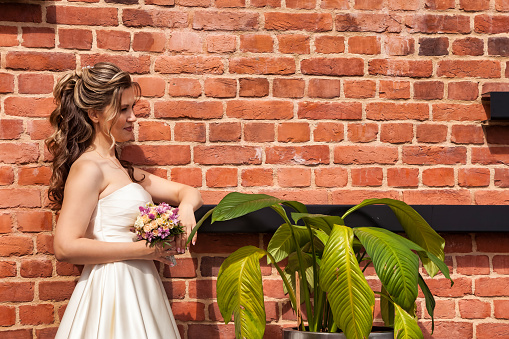 French bride looking away with bouquet in white dress at background of decorated brick wall. Bridal portrait of pretty fiancee outdoors. Happy marriage and family creation concept. Copy text space