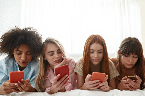 Young multiethnic girlfriends typing and browsing smartphones on bed during girlish pajama party at home. Black and caucasian zoomer girls resting together. Friendship. Entertainment and leisure