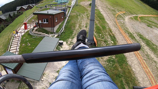 Girl rides or climbs on lift or cable car up mountain. Top view. Girl's legs in blue jeans and black sneaker stand on footrest, movement of cable car seat over path on which person is walking close-up