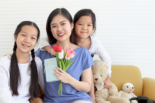 Two daughter girls give tulip flower and present gift box for smiling mother while hug mom with love at living room, warm love in family. Happy mother day, children have special time with mom.