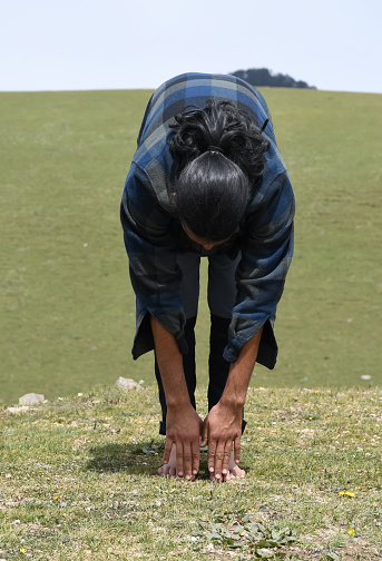 A long haired young man doing warm up by Uttanasana Yoga Pose (Standing Toe Touch) stretch exercise in the nature.