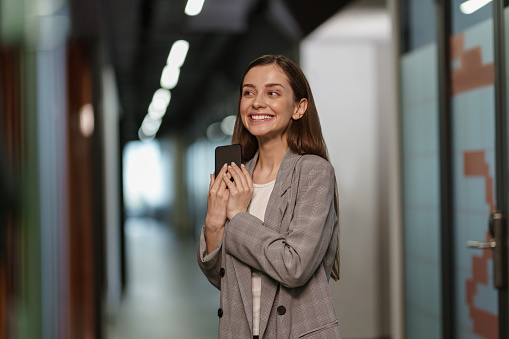 Portrait of attractive business woman with phone standing in modern coworking and looking away