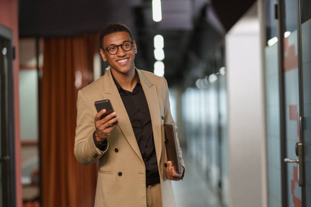 istock Handsome smiling african businessman using phone while standing in modern coworking 1418728424