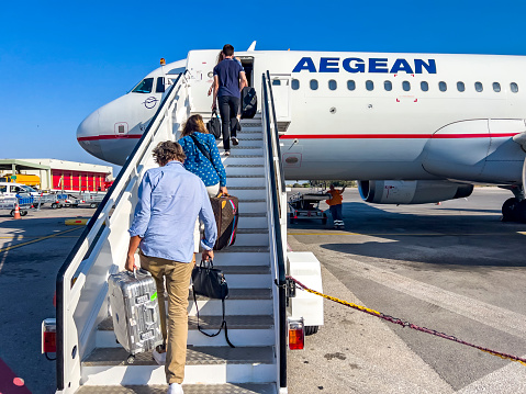 Athens, Greece - July 26, 2022: People climbing on the airplane jet ladder with their luggage at Athens International Airport Eleftherios Venizelos. Going on vacation with Aegean airlines. Adults walking up the stairs towards the plane. Company logo and brand for advertisement. Accessibility for people. Travel theme background on sunny day