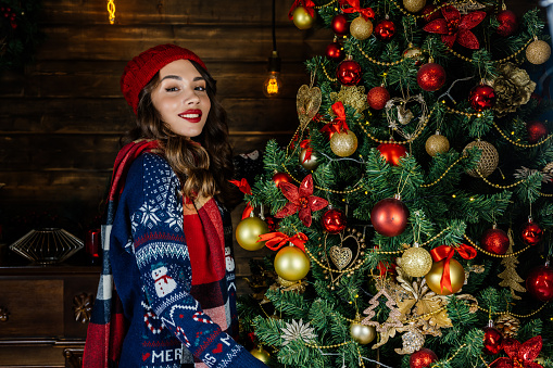 A woman in a red hat and scarf looks joyfully. A beautiful brunette stands by the fireplace against the backdrop of a Christmas tree and Christmas decorations.