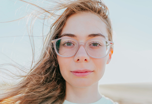 Bright outdoor portrait of caucasian teenager girl with glasses. Long blond hair blowing in the wind. Young face close up.