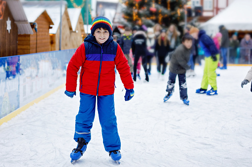 Happy little kid boy in colorful warm clothes skating on a rink of Christmas market or fair. Healthy child having fun on ice skate. Lot of people celebrating holiday and having active winter leisure.